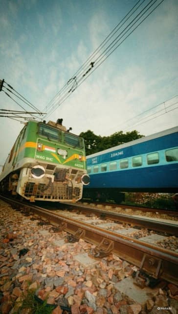A green electric locomotive crossing with a blue train behind